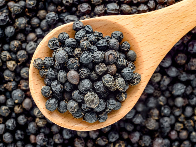 Wooden spoon filled with black peppercorns on a background of black peppercorns.