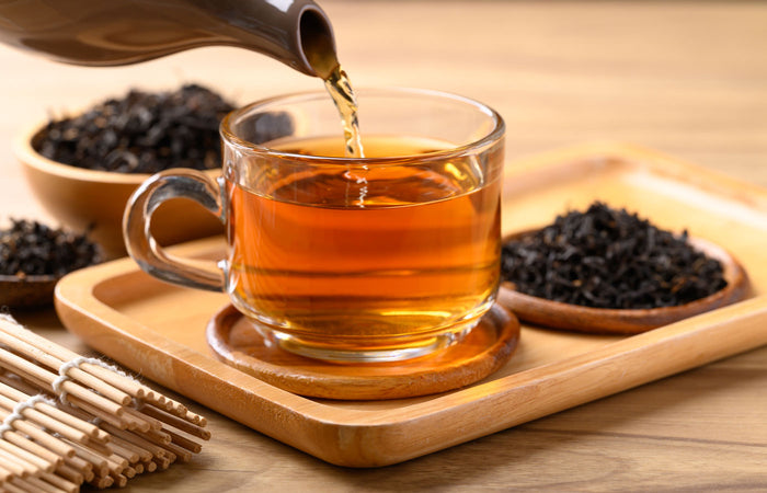 Tea being poured into a glass cup with tea leaves and a wooden tray in the background.