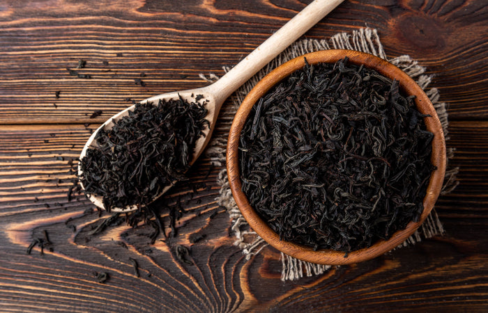 Wooden spoon and bowl filled with dark dried black tea leaves on a wooden surface