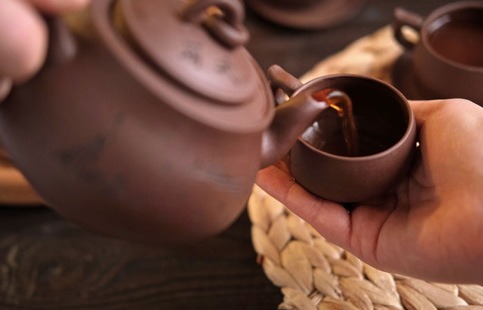 Person pouring tea from a purple clay teapot into a matching cup.