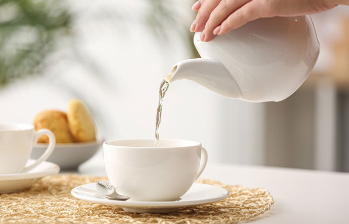 Tea being poured from a white teapot into a white cup on a woven mat with a blurred background.