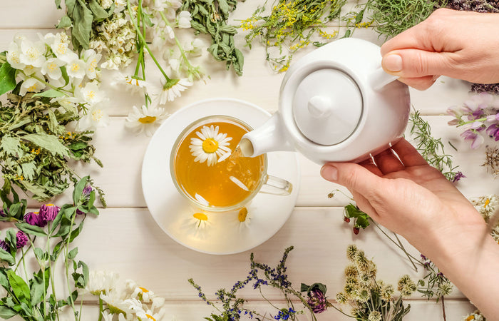 Person pouring tea into a cup with chamomile flowers and herbs on a wooden surface