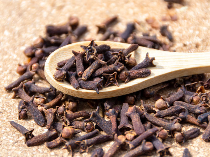 cloves on a wooden spoon with a pile of herbs on a sandy background