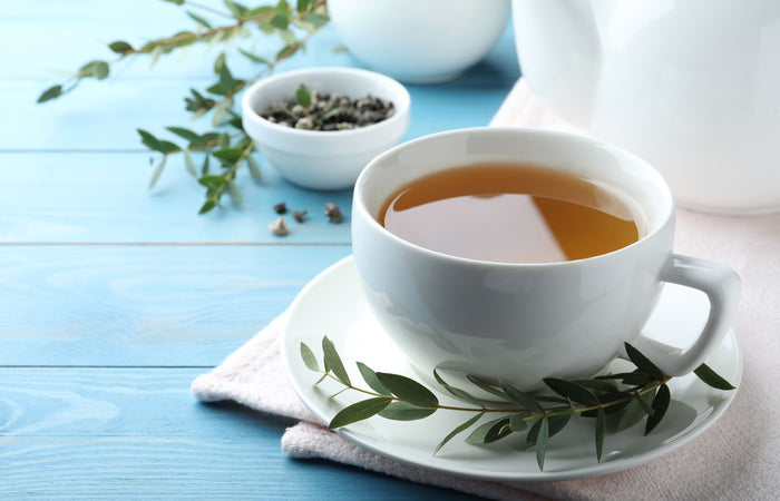 White teacup with tea on a saucer, surrounded by tea leaves and a teapot on a light blue wooden surface.