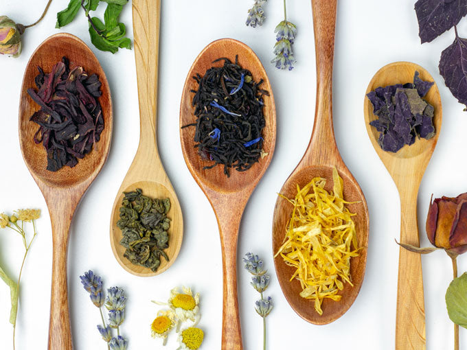 Wooden spoons with various types of tea leaves and flowers on a white background