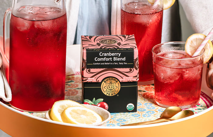 Cranberry comfort blend tea packaging with a pitcher and glasses of red liquid on a tray.