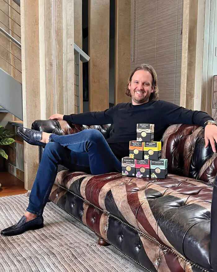 John Boyd sitting on a patterned sofa with a stack of Buddha Teas tea boxes in front of him.