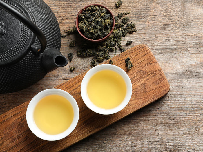 Tea-making setup with black teapot, two white cups, and a small bowl of oolong tea leaves on a wooden board.
