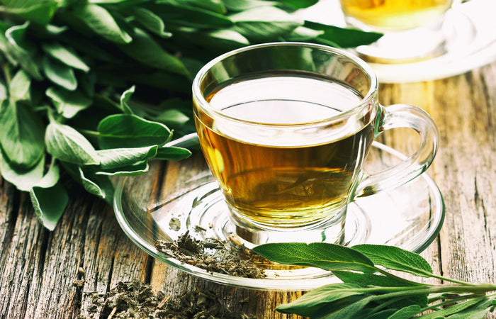 Clear glass cup filled with sage tea on a wooden surface with green sage leaves.