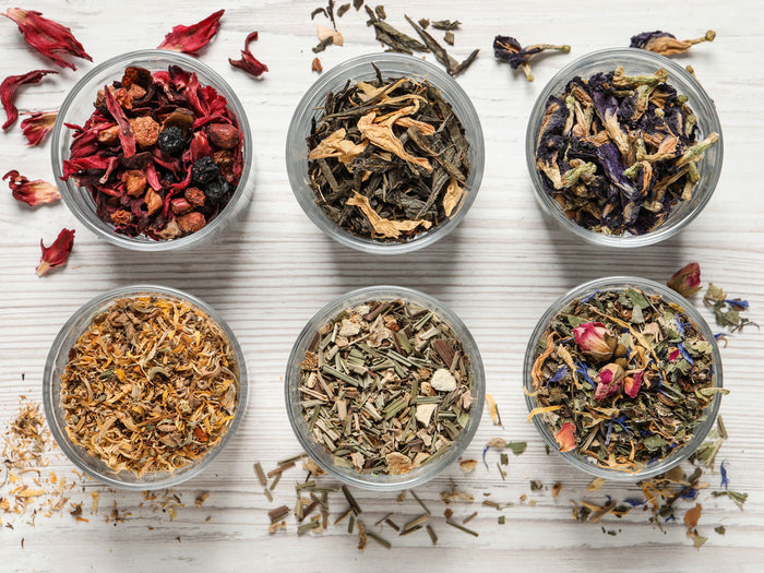 Six small bowls filled with various types of dried tea leaves on a light wooden surface.
