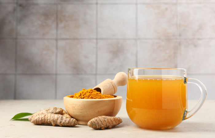 Glass mug of orange juice with a wooden bowl of turmeric and turmeric roots on a light surface.